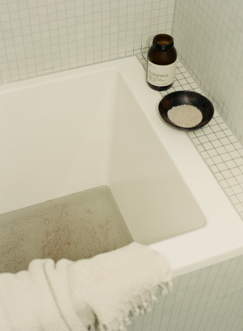 Enlighten bath salts bottle on tiled bath side with salts in bath. Waffle towel and small dish with salts